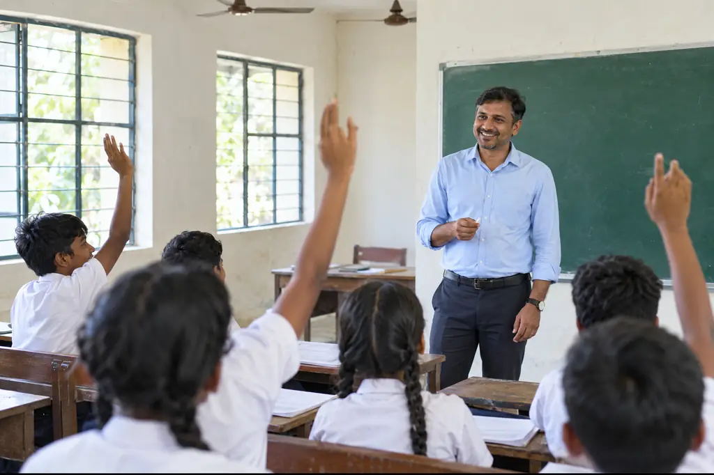 A teacher smiles as students raise their hands in a government school classroom